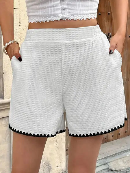 Outdoor look of a woman modeling high-rise white shorts with black scallop trim and pockets, styled with a vintage-inspired white crop top—perfect for warm weather chic.